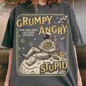 May include: Dark gray t-shirt featuring a retro-style illustration of an astronaut on a moon, with the words "GRUMPY ANGRY STUPID". The design includes a rocket, tent, and the question "HOW LONG SINCE LAST SLEEP QUESTION?"