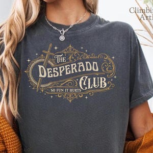 Peut inclure: T-shirt gris foncé avec l'inscription "The Desperado Club" dans un design décoratif, avec une épée et la phrase "So Fun It Hurts". Un vêtement confortable et décontracté.