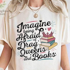 Op de afbeelding: Crèmekleurig T-shirt met de tekst "Imagine being Afraid of Drag Queens and Books" in een zwarte, gestileerde letter. Het ontwerp bevat een roze kroon, een roze hart en een stapel boeken.