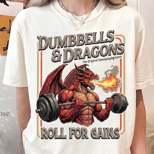 Op de afbeelding: Crèmekleurig T-shirt met een afbeelding van een gespierde rode draak die een halter optilt, met de tekst "DUMBBELLS & DRAGONS" en "ROLL FOR GAINS". Het shirt heeft ook de tekst "The Original Swoleplaying Game".