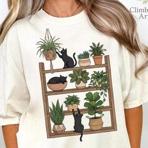 May include: Cream-colored t-shirt featuring a graphic of a wooden plant shelf with various potted plants and three black cats. The shelf has three levels, with the cats interacting with the plants. The text "ClimbingRose Artisan" is on the upper right.