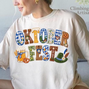 Puede incluir: Una camiseta de color crema con el texto OKTOBERFEST en letras grandes y decorativas. Las letras están rellenas con patrones que incluyen cuadros vichy azules y blancos, pretzels, jarras de cerveza, sombreros verdes con plumas y motivos florales.