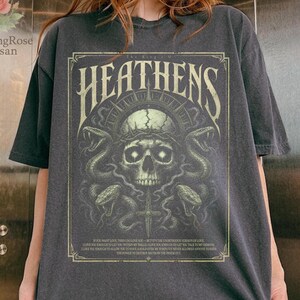 Puede incluir: Camiseta gris oscuro con un gráfico detallado de una calavera, serpientes y el texto "HEATENS". El diseño está enmarcado con un borde decorativo. La camiseta tiene mangas cortas y un corte holgado.