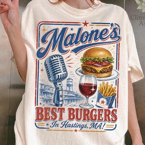May include: Cream-colored t-shirt featuring a vintage-style graphic for "Malone's Best Burgers." The design includes a burger, fries, wine, and a microphone. Text reads "In Hastings, MA!"