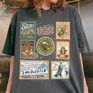 Puede incluir: Camiseta gris oscuro con diseños de sellos postales con texto e ilustraciones de una novela de fantasía. Los sellos representan un anillo, personajes y paisajes. La camiseta también tiene el logotipo de ClimbingRose Artisan.