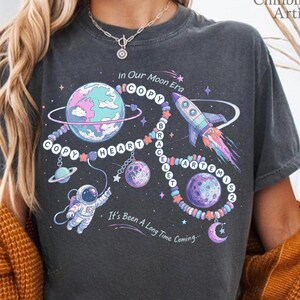 May include: Dark gray t-shirt with a colorful space-themed graphic. The design includes planets, a rocket, an astronaut, and the text "In Our Moon Era." The phrase "It's Been A Long Time Coming" is also featured.