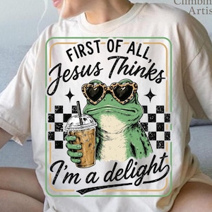 May include: A cream-colored short-sleeve t-shirt featuring a graphic of a green frog wearing heart-shaped sunglasses and holding a coffee cup. The text reads: First of all, Jesus Thinks I'm a delight. The design includes a black and white checkerboard pattern.