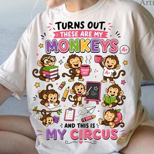 Könnte beinhalten: Ein cremefarbenes T-Shirt mit einer bunten Grafik von mehreren Zeichentrickaffen bei schulischen Aktivitäten. Der Text lautet "Turns out, these are my monkeys and this is my circus" und zeigt Illustrationen von Büchern, Stiften und einer Tafel.