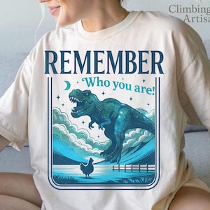 Puede incluir: Camiseta beige con un diseño de dinosaurio turquesa y la frase en azul marino "REMEMBER Who you are!". El diseño incluye una luna, estrellas, nubes y una gallina. Prenda de vestir informal.