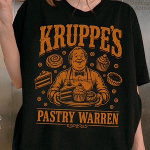 May include: A black t-shirt featuring an orange graphic print of a person wearing an apron and a bowtie, holding a tray of pastries. The text reads KRUPPE'S PASTRY WARREN, surrounded by illustrations of various cakes, cupcakes, and cinnamon rolls.