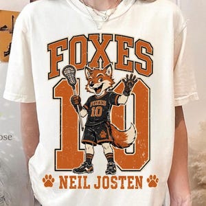 May include: Cream-colored t-shirt featuring a cartoon fox lacrosse player with the word "FOXES" above the number "10" in orange. The text "NEIL JOSTEN" is below the number. The fox is wearing a black and orange uniform.