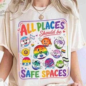 Könnte beinhalten: Cremefarbenes T-Shirt mit einem farbenfrohen Aufdruck mit der Aufschrift "ALL PLACES Should be SAFE SPACES". Die Grafik zeigt verschiedene emoji-ähnliche Charaktere mit verschiedenen Pride-Flaggen. Das Shirt hat kurze Ärmel und einen Rundhalsausschnitt.
