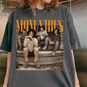 May include: Dark gray t-shirt with the words "MOM VIBES" in orange block letters. The shirt features a vintage-style image of four people sitting on a porch. The shirt is a relaxed fit.