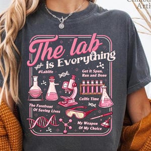 May include: Dark grey t-shirt with a pink and white graphic that reads "The lab is Everything." The graphic includes lab equipment illustrations and the phrases "#Lablife," "Get It Spun, Run and Done," and "Cellfe Time."