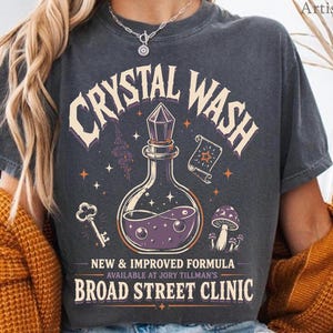 May include: Dark gray t-shirt with the words "CRYSTAL WASH" above a potion bottle graphic. Below the graphic, the text reads "NEW & IMPROVED FORMULA" and "BROAD STREET CLINIC". The shirt has a vintage, slightly faded look.