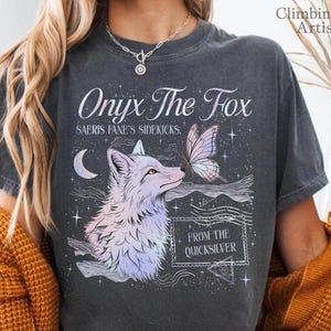Può includere: T-shirt grigio scuro con la scritta "Onyx The Fox" e "Saeris Fane's Sidekicks." Il design presenta una volpe, una farfalla, una luna crescente e le parole "From The Quicksilver". La maglietta ha un aspetto vintage.