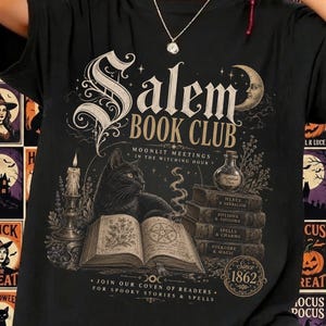 Könnte beinhalten: Ein schwarzes T-Shirt mit einem Grafikdruck, der den Text "Salem Book Club" und "Since 1862" enthält. Das Design zeigt eine schwarze Katze auf einem offenen Buch, einen Stapel Bücher mit verschiedenen magischen Themen, eine Kerze und eine Mondsichel.