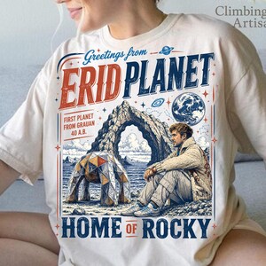 May include: A cream-colored t-shirt featuring a graphic print with the text Greetings from ERID PLANET HOME OF ROCKY. The design includes a person sitting near a geometric dome structure, a large rock arch, a planet, and stars in blue and red ink.