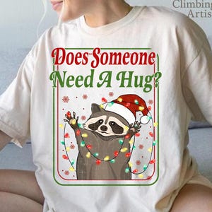 May include: Beige t-shirt featuring a raccoon wearing a Santa hat and wrapped in Christmas lights. The text "Does Someone Need A Hug?" is displayed in red and green. A festive design for the holiday season.