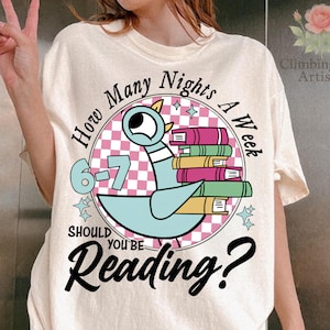 May include: Cream-colored t-shirt featuring a cartoon pigeon holding a stack of books. The design includes the text "How Many Nights A Week Should You Be Reading?" and the numbers "6-7". The shirt has a round neck and short sleeves.