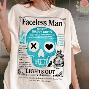 Può includere: T-shirt color crema con una grafica raffigurante un teschio turchese con una croce e un cuore al posto degli occhi, e le parole "Faceless Man" e "Lights Out". La maglietta presenta vari altri elementi testuali e illustrazioni.