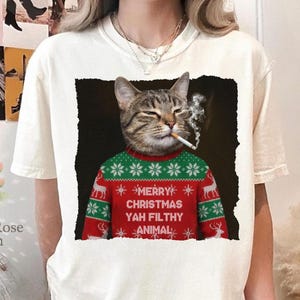 May include: Cream-colored t-shirt featuring a cat wearing a Christmas sweater and smoking a cigarette. The red and green sweater has a snowflake and reindeer pattern, with the text "MERRY CHRISTMAS YAH FILTHY ANIMAL".