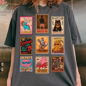 May include: Dark gray t-shirt featuring nine colorful postage stamp-style graphics. The graphics include cartoon characters, animals, and text such as "MONGO IS APPALLED", "YOU WILL NOT BREAK ME", and "PRINCESS DONUT". The shirt has a relaxed fit.