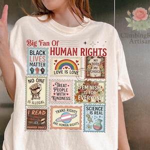 May include: Cream-colored t-shirt with a graphic of postage stamp designs. The stamps feature phrases like "Black Lives Matter," "Love is Love," and "Trans Rights are Human Rights." The shirt also includes the text "Big Fan Of Human Rights."