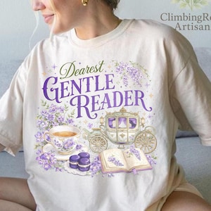 May include: Cream-colored t-shirt featuring a whimsical design with the text "Dearest Gentle Reader" in purple script. The graphic includes a teacup, carriage, book, and macarons, all in shades of purple and white. The shirt is a relaxed fit.