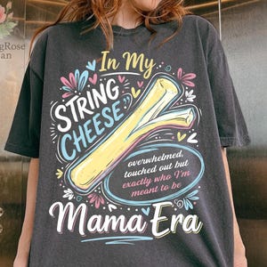 May include: Dark grey t-shirt with a playful design featuring a string cheese illustration. The text reads "In My String Cheese Mama Era" with additional text: "overwhelmed, touched out but exactly who I'm meant to be."