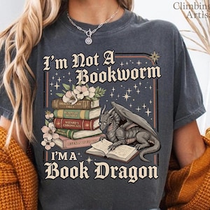 Może przedstawiać: Ciemnoszara koszulka z napisem "I'm Not A Bookworm, I'm A Book Dragon." Grafika przedstawia smoka czytającego książkę, ze stosem książek i kwiatowymi akcentami. Koszulka jest w wygodnym, swobodnym stylu.