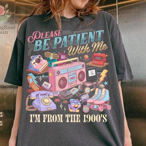 Puede incluir: Camiseta gris oscuro con un gráfico colorido que presenta una radio rosa, patines y otros artículos retro. El texto dice "PLEASE BE PATIENT With Me I'M FROM THE 1900'S".