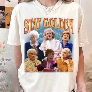 May include: Cream-colored t-shirt featuring a graphic of the Golden Girls cast with the words "Stay Golden" in orange and yellow. The shirt is a short-sleeved crew neck.