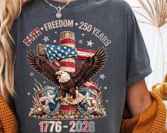 Puede incluir: Camiseta gris oscuro con un diseño patriótico. Presenta una cruz con la bandera estadounidense, un águila con las alas extendidas y el texto "Faith, Freedom, 250 Years, 1776-2026". Dos escudos con estrellas en la parte inferior.
