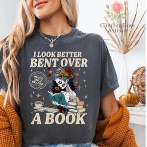 May include: Dark gray t-shirt with the text "I LOOK BETTER BENT OVER A BOOK." The shirt features a graphic of a skeleton reading a book, with floral and butterfly accents. A coffee cup and the words "Proud Smut Reader" are also visible.