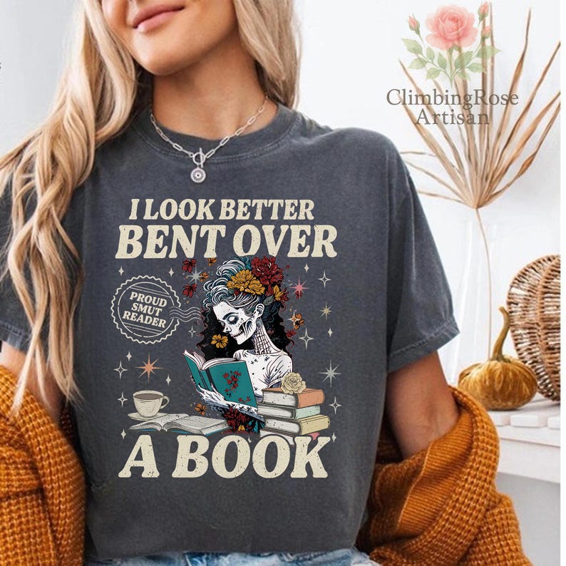 May include: Dark gray t-shirt with the text "I LOOK BETTER BENT OVER A BOOK." The shirt features a graphic of a skeleton reading a book, with floral and butterfly accents. A coffee cup and the words "Proud Smut Reader" are also visible.