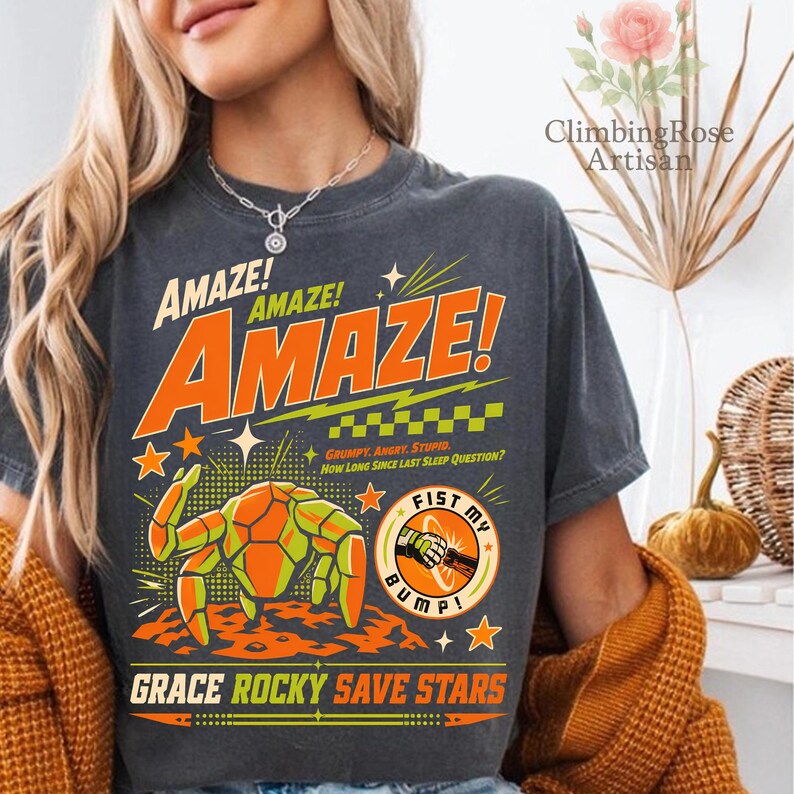 K&ouml;nnte beinhalten: Dunkelgraues T-Shirt mit dem Wort "AMAZE" in gro&szlig;en orangefarbenen Buchstaben. Das Shirt zeigt eine Grafik einer krabbenartigen Kreatur und den Text "FIST MY BUMP!" und "GRACE ROCKY SAVE STARS". Das Shirt hat auch den Text "GRUMPY. ANGRY. STUPID. How Long Since Last Sleep Question?"