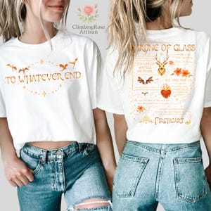 May include: White t-shirts with gold-colored graphics. One shirt reads "TO WHATEVER END" with a decorative border and flying creatures. The other shirt features text that reads "THRONE OF GLASS" and "FIREHEART" with other decorative elements.