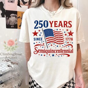 May include: Cream-colored t-shirt with a patriotic design. The design features the text "250 YEARS SINCE 1776 Semiquincentennial" in blue and red, along with an American flag and stars. The shirt is a casual style.