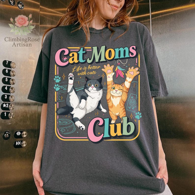 Op de afbeelding: Donkergrijs t-shirt met een kleurrijke afbeelding met de tekst "Cat Moms Club". Het ontwerp toont twee cartoonkatten, een zwart-witte, de andere oranje, met de tekst "Life is better with cats."