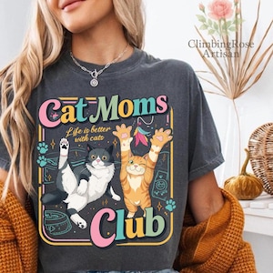 Op de afbeelding: Donkergrijs t-shirt met een kleurrijke afbeelding met de tekst "Cat Moms Club". Het ontwerp toont twee cartoonkatten, een grijze en witte, de andere oranje en witte. De tekst "Life is better with cats" is ook opgenomen.