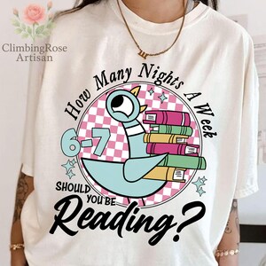 May include: Cream-colored t-shirt with a graphic featuring a cartoon pigeon carrying a stack of books. The design includes the text "How Many Nights A Week Should You Be Reading?" and the numbers "6-7".
