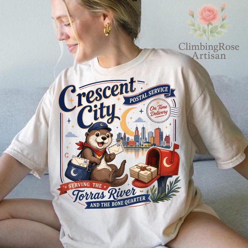 May include: Cream-colored t-shirt featuring a whimsical illustration of an otter in a postal uniform. The design includes a mailbox, cityscape, and the text "Crescent City Postal Service". The shirt also has the text "Serving the Torras River and the Bone Quarter".