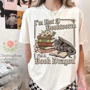 Pode incluir: Camiseta creme com o texto "I'm Not A Bookworm, I'm A Book Dragon". O gr&aacute;fico apresenta uma pilha de livros, flores e um drag&atilde;o lendo um livro. A camisa &eacute; de estilo casual.