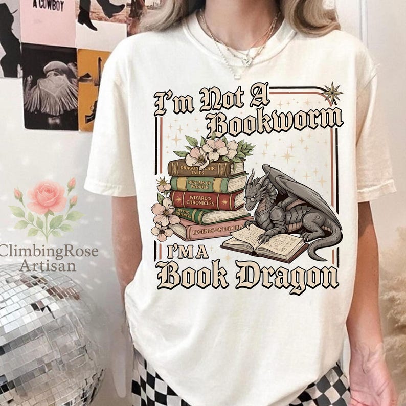 Pode incluir: Camiseta creme com o texto "I'm Not A Bookworm, I'm A Book Dragon". O gr&aacute;fico apresenta uma pilha de livros, flores e um drag&atilde;o lendo um livro. A camisa &eacute; de estilo casual.