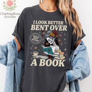 May include: Dark gray t-shirt with the text "I LOOK BETTER BENT OVER A BOOK." The graphic features a skeleton reading a book, adorned with flowers. A coffee cup and books are also depicted.