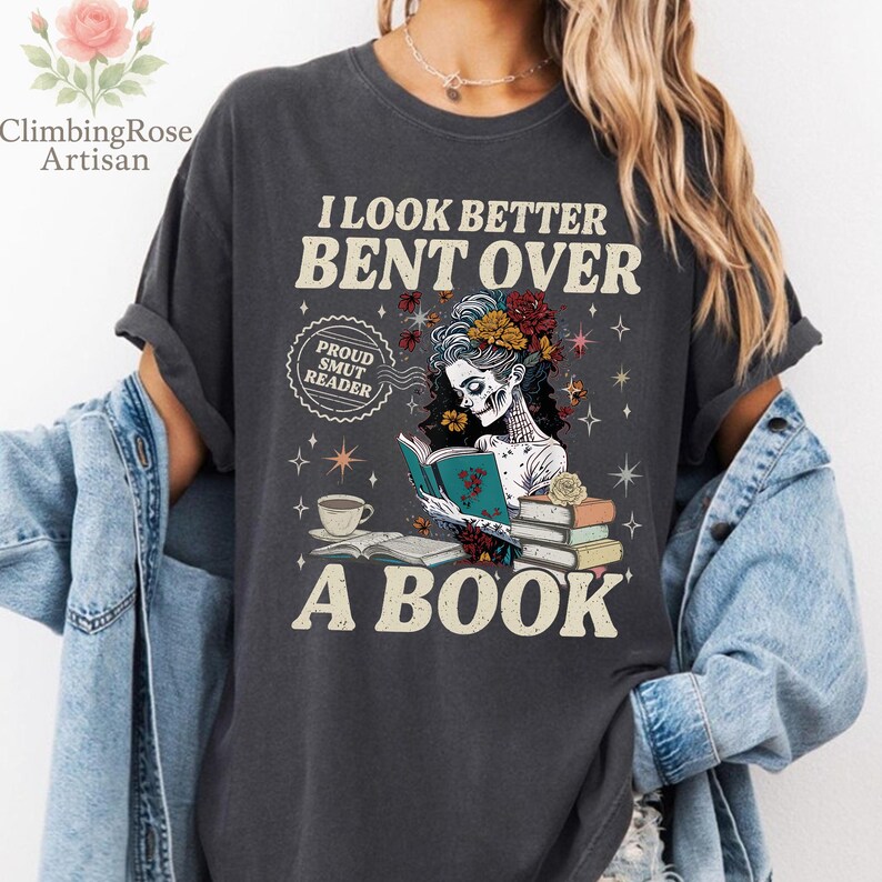 May include: Dark gray t-shirt with the text "I LOOK BETTER BENT OVER A BOOK." The graphic features a skeleton reading a book, adorned with flowers. A coffee cup and books are also depicted.