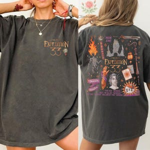 May include: Dark gray t-shirt with "EXPEDITION 33" in orange text on the front. The back features a detailed graphic with text, a flame, and various illustrations. The shirt is a comfortable, oversized fit.