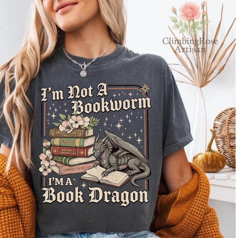 Pode incluir: T-shirt cinzenta escura com o texto "I'm Not A Bookworm, I'm A Book Dragon." O gr&aacute;fico apresenta um drag&atilde;o lendo um livro, com uma pilha de livros e detalhes florais. A camisa &eacute; um estilo confort&aacute;vel e casual.