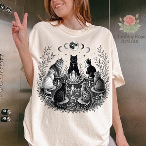May include: A cream-colored t-shirt featuring a black ink graphic of seven cats sitting on tree stumps in a circle around a central crystal. The design includes a moon phase cycle above the cats, surrounded by botanical elements and mushrooms.